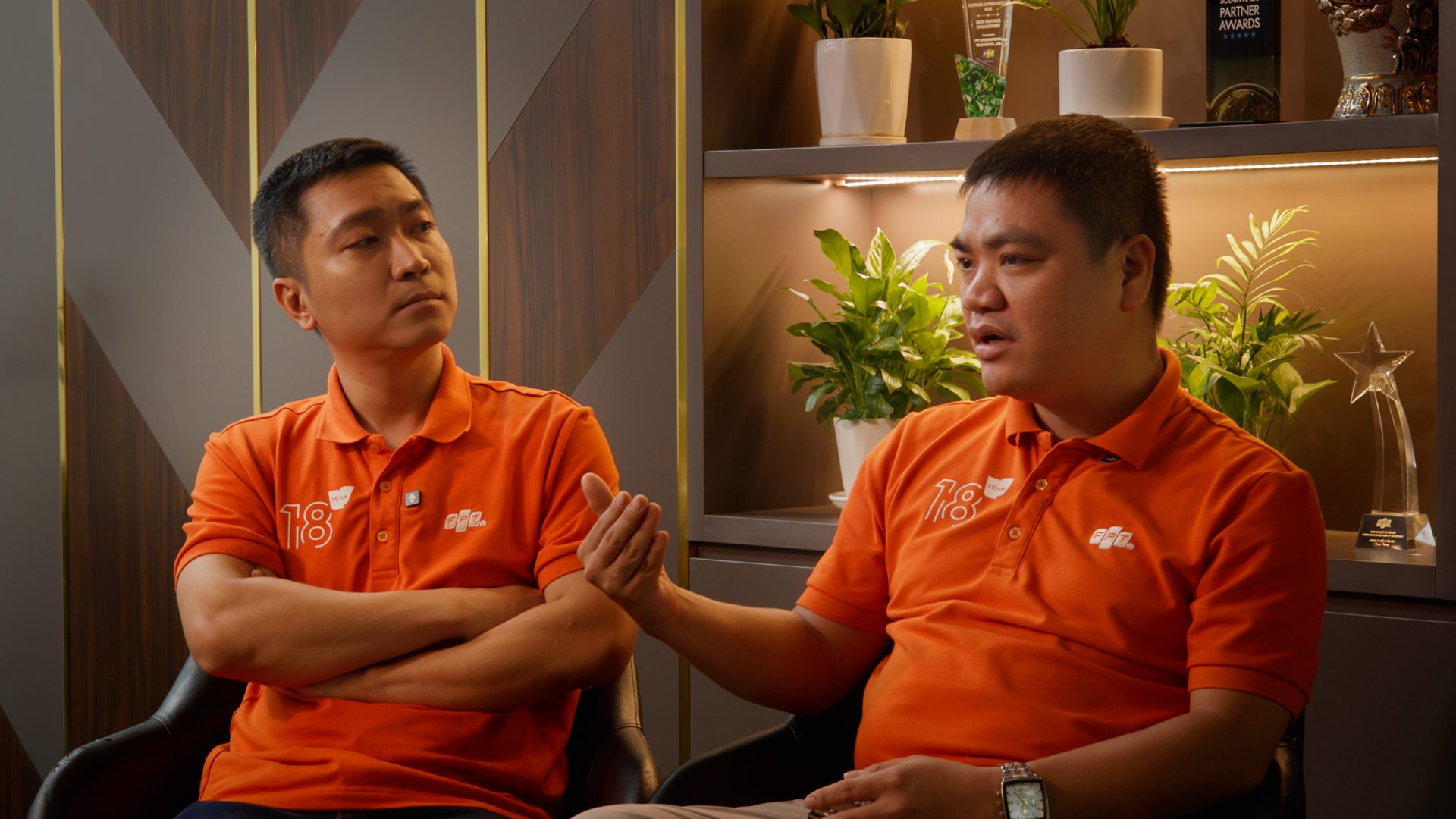 Two FPT Telecom staff in orange polos seated for a paired interview in front of an awards shelf